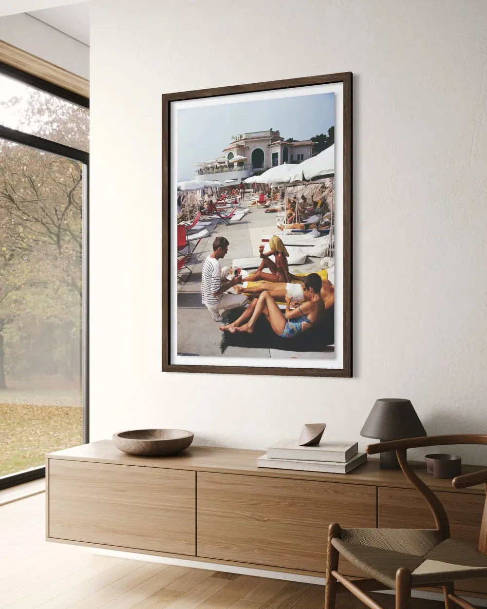 A framed vertical art print depicting a sunny beach scene with people lounging under white umbrellas, displayed on a white wall above a light wood console table in a modern interior setting.