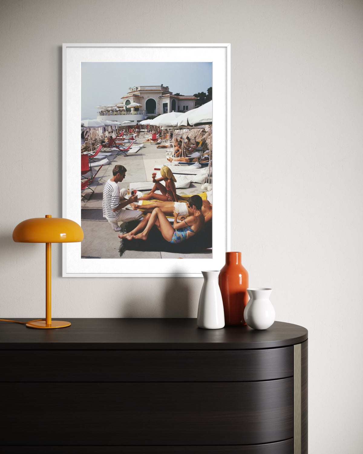 A framed art print depicting a sunny beach scene with people lounging under umbrellas, displayed on a light gray wall above a dark wood console table with decorative vases and a yellow lamp.