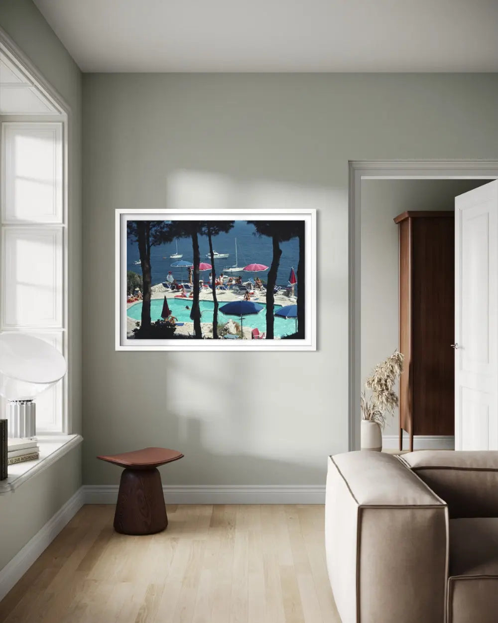A framed art print depicting a vibrant outdoor swimming pool scene with palm trees and beach umbrellas, displayed on a light sage green wall in a modern living room setting.