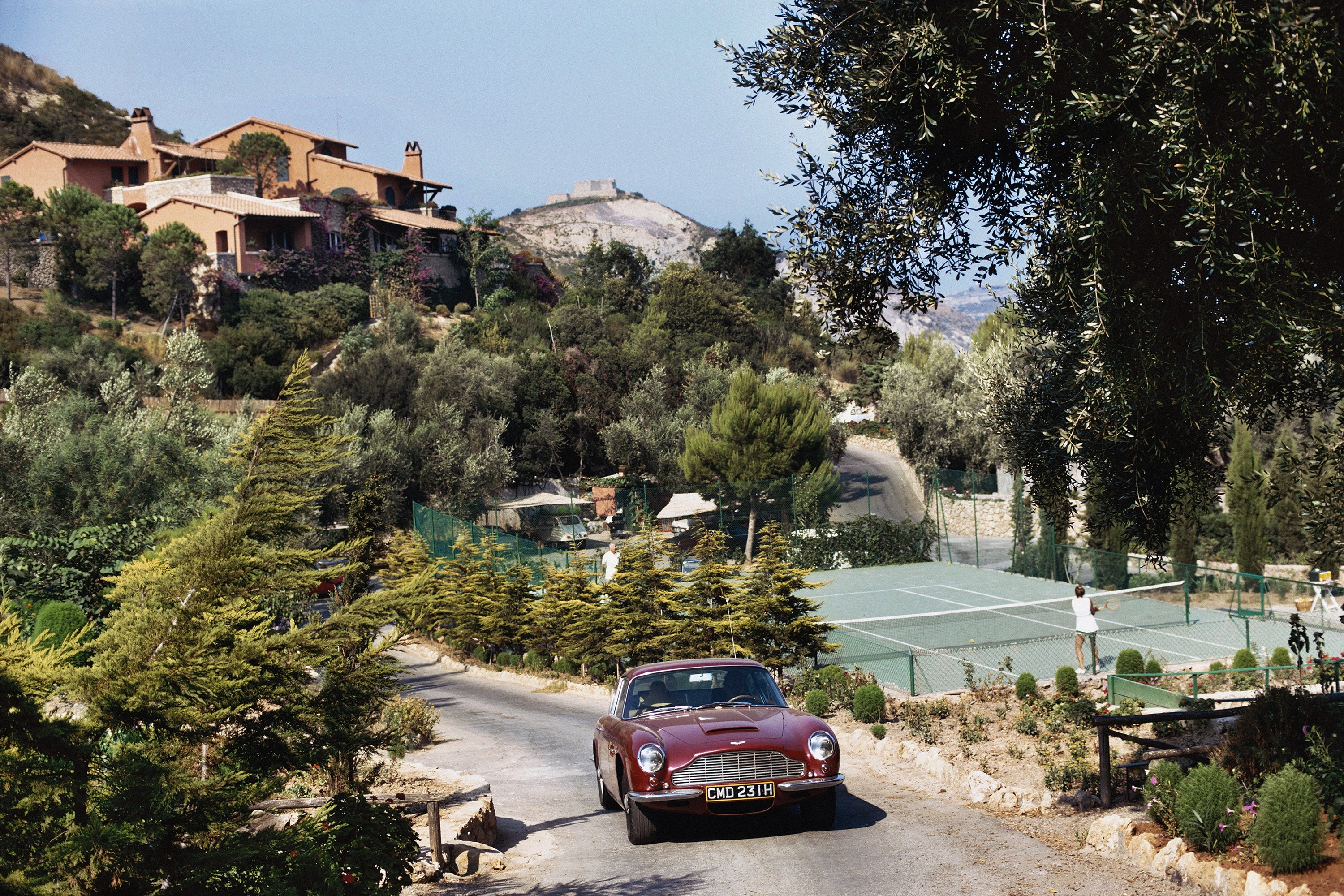 A vintage red Aston Martin sports car drives down a winding road in a scenic, upscale outdoor setting with tennis courts, trees, and hillside villas in the background.