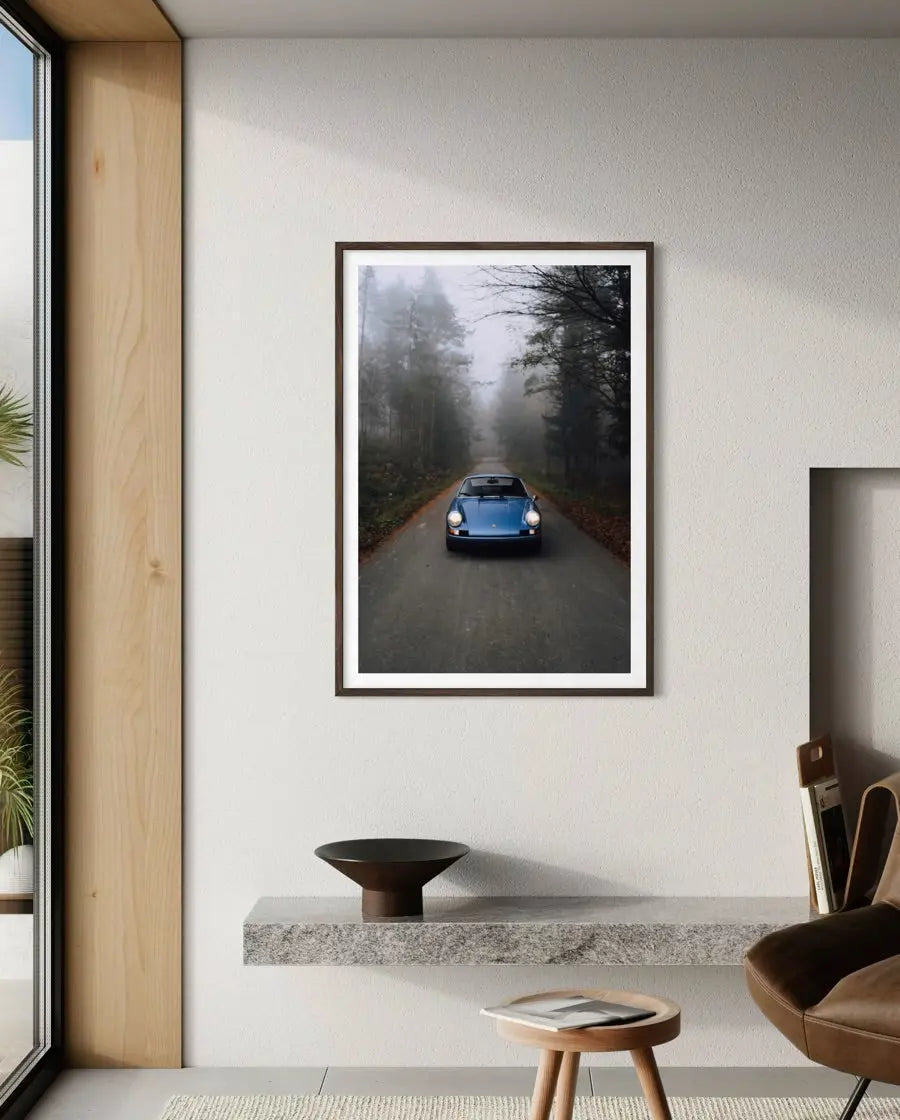 A framed vertical art print of a classic blue car driving down a foggy forest road, displayed on a light gray wall in a modern interior setting.