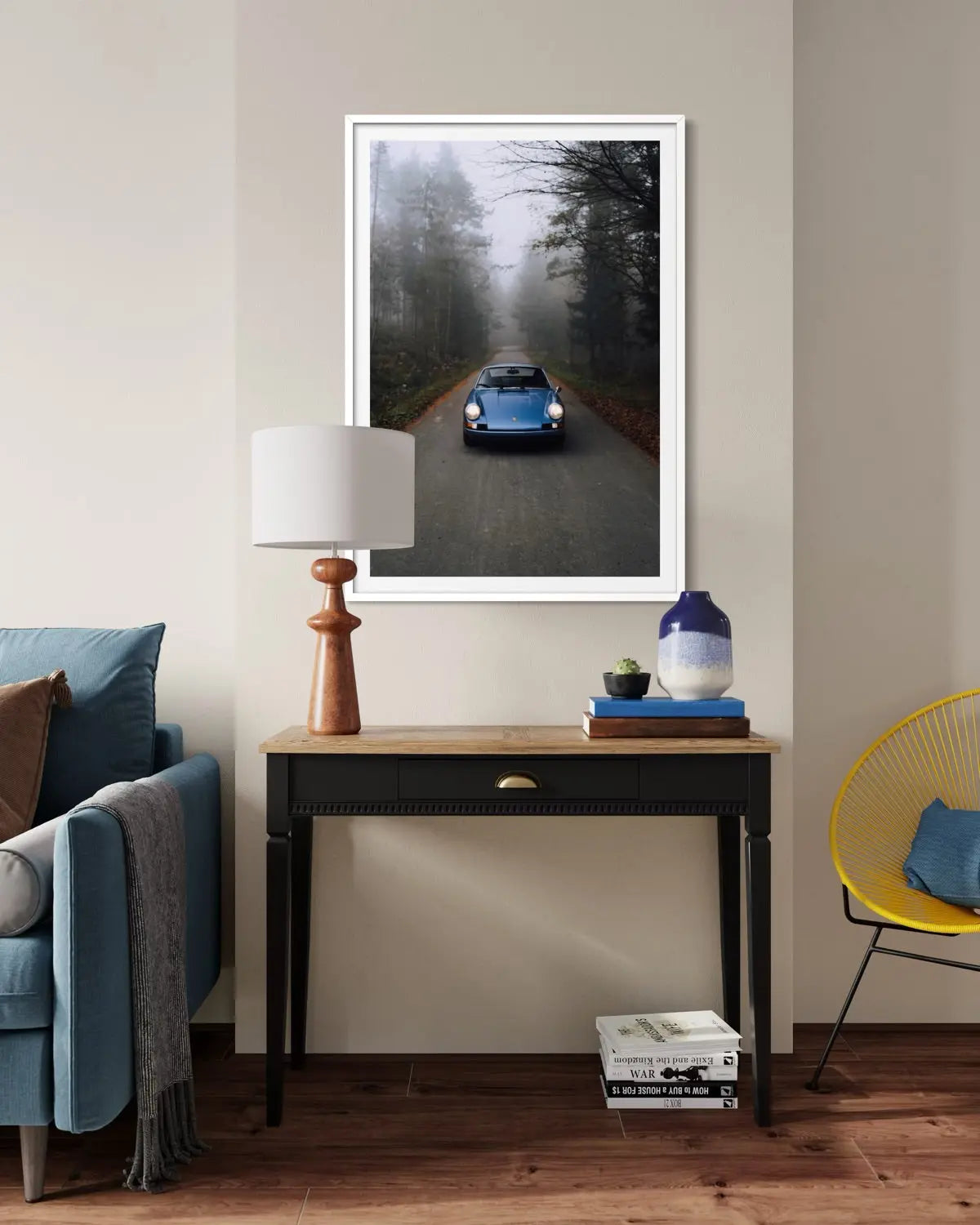A framed vertical art print of a blue vintage car driving down a foggy forest road, displayed on a beige wall above a console table in a styled living room setting.