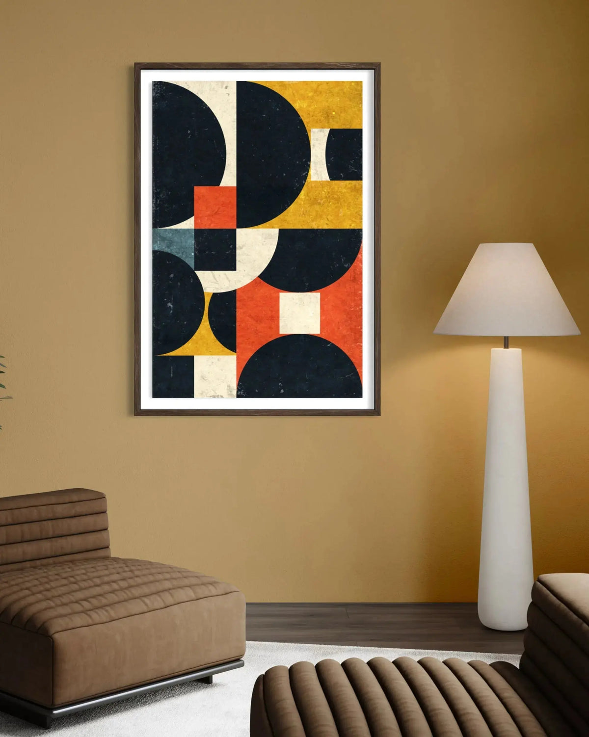 A framed abstract geometric wall art print hangs on a warm mustard-yellow wall in a modern living room setting, featuring bold shapes in navy, mustard yellow, terracotta, cream, and orange. The print is shown alongside a brown leather ottoman, a white floor lamp, and a dark wood floor.