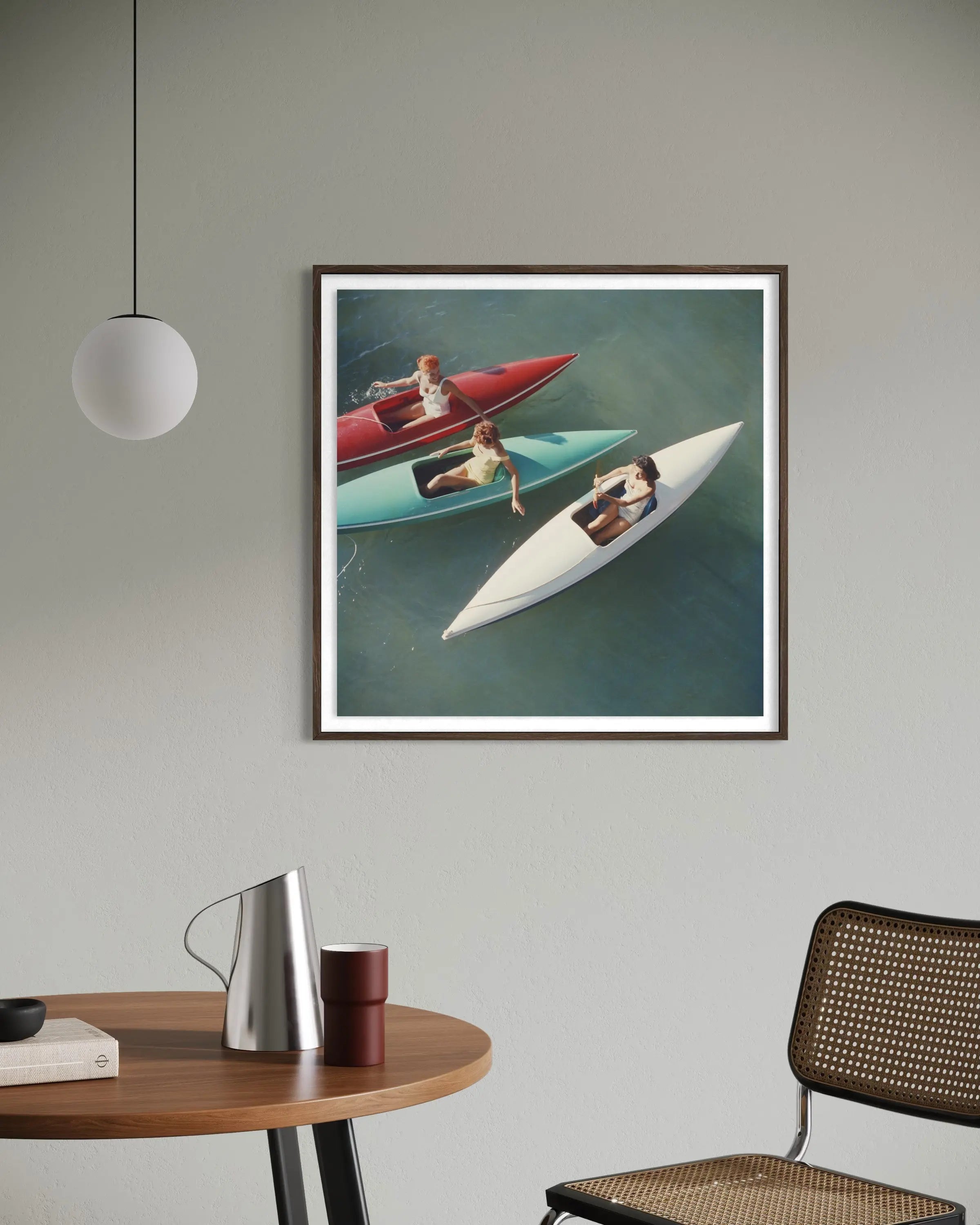A framed art print depicting three people in small boats on blue-green water, hanging on a light gray wall in a modern room setting with a wooden table, chair, and pendant light visible.