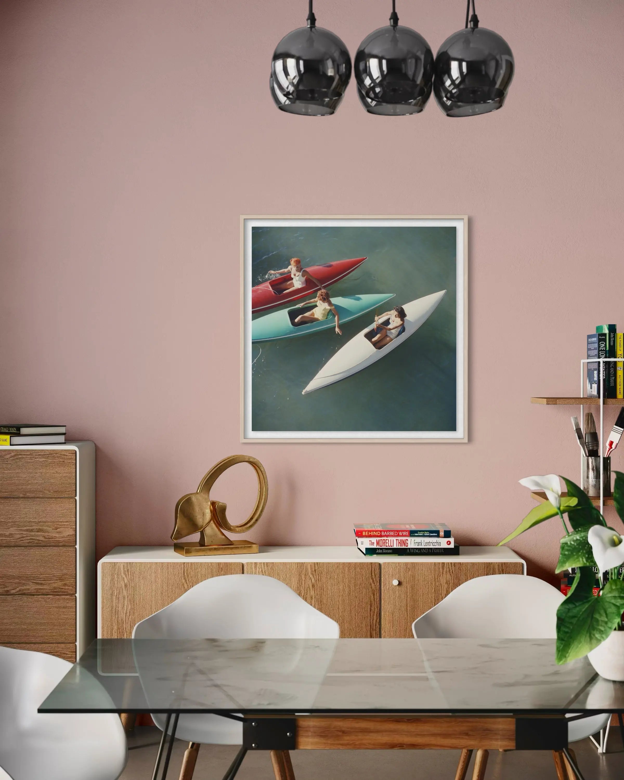 A framed art print depicting two people in colorful rowboats on a teal lake, displayed on a dusty pink wall in a modern dining room setting above a wooden sideboard and dining table.