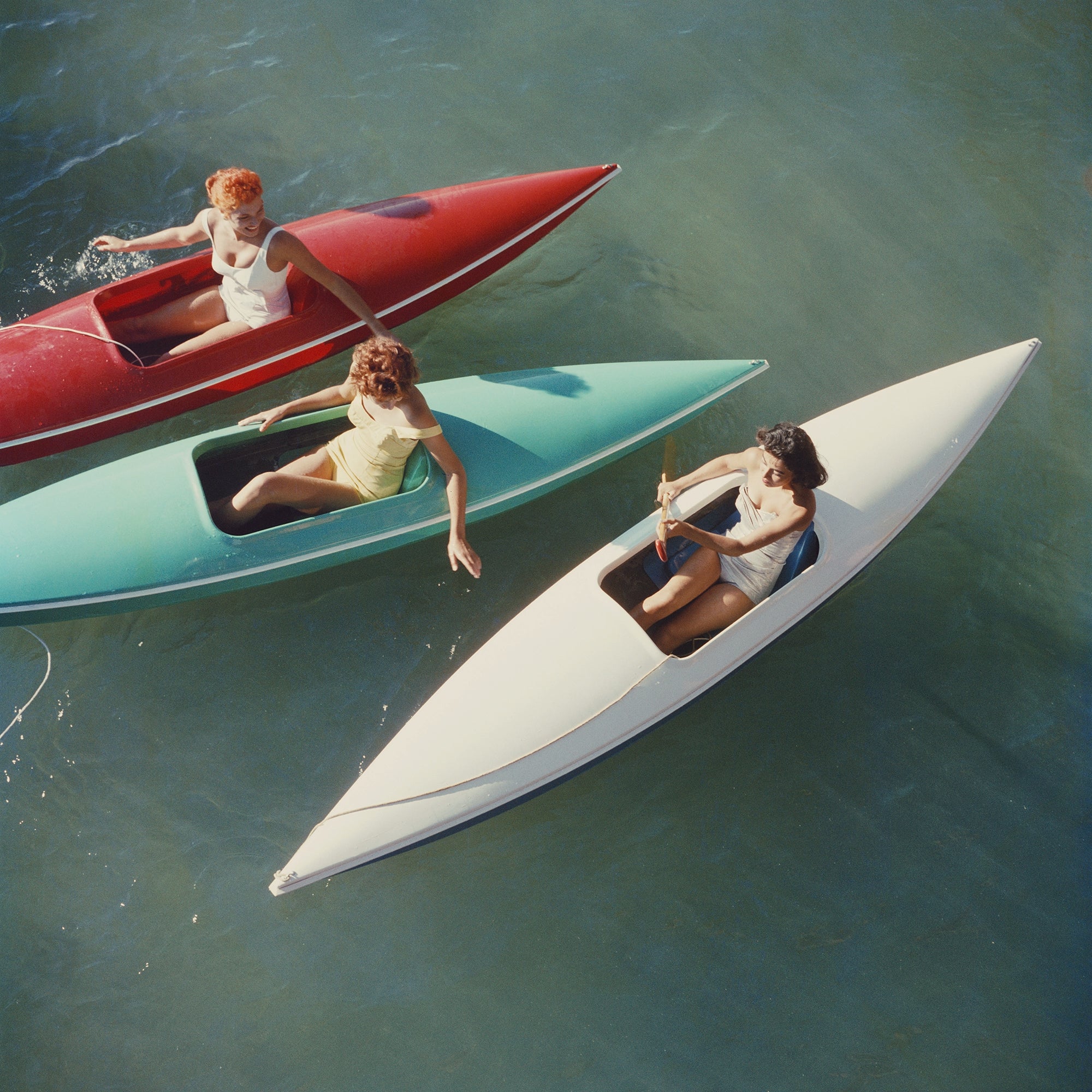 Three women sit in narrow, boat-like watercraft on open water. One craft is red, one is aqua/turquoise, and one is white, arranged diagonally across blue-green water.