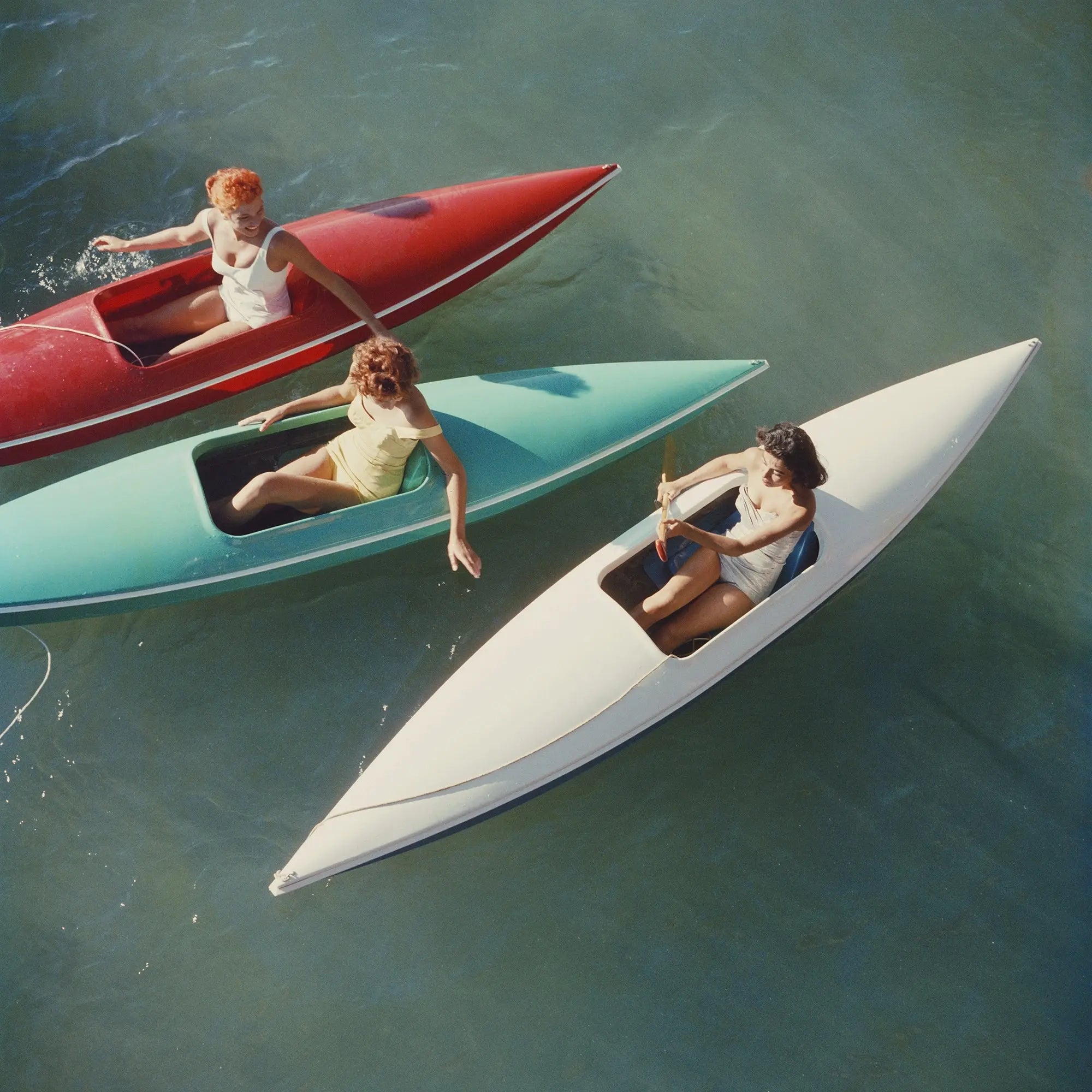 Three women sit in narrow, boat-like watercraft on open water. One craft is red, one is aqua/turquoise, and one is white, arranged diagonally across blue-green water.