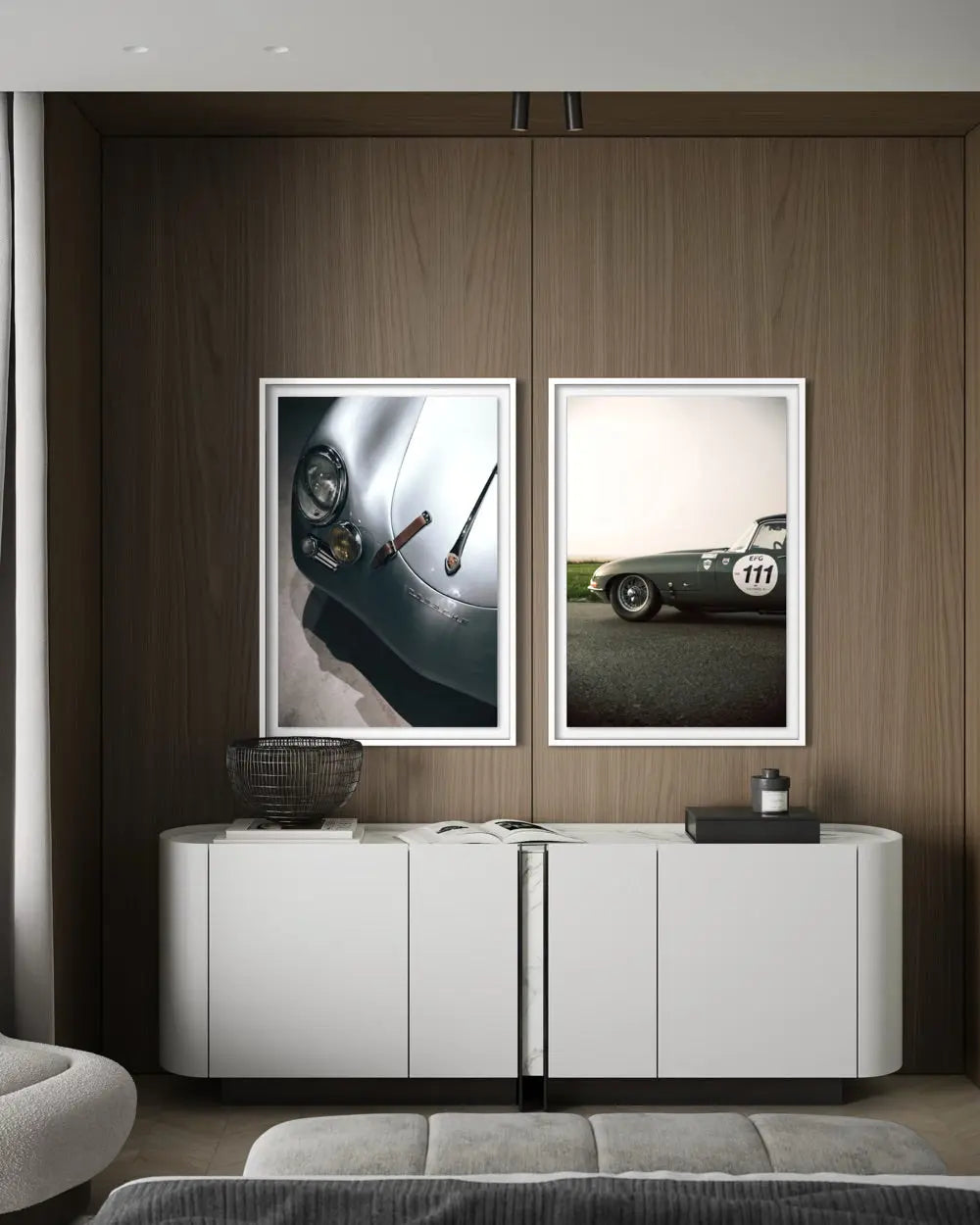 Two framed vertical art prints depicting classic race cars, displayed side-by-side on a dark wood wall above a white sideboard in a modern living room setting.