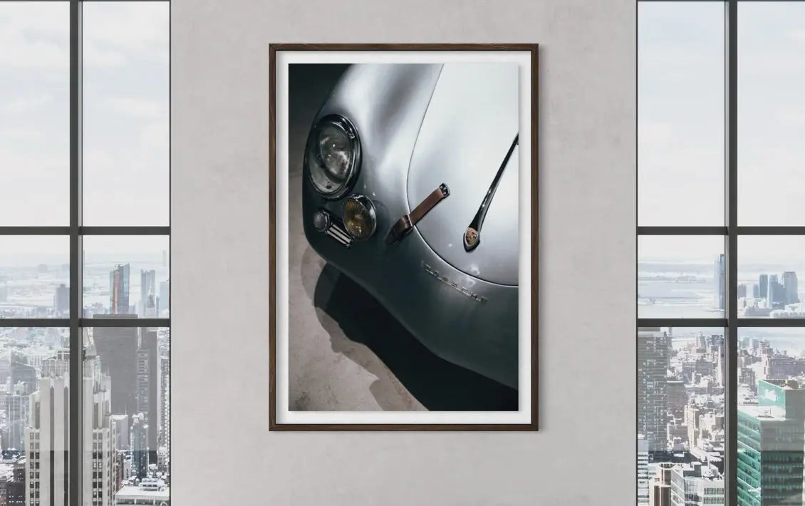 A framed art print depicting a close-up of a silver classic car with a cigar, displayed on a light gray wall between two large windows showing a city skyline.