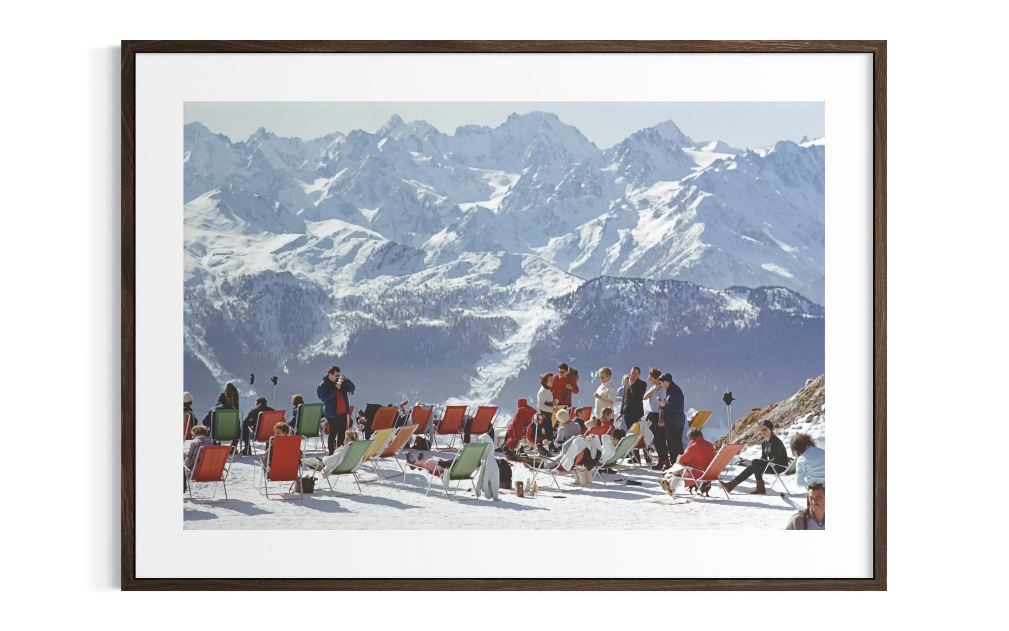 A framed photographic print of a snowy mountain landscape with people sitting in red and green beach chairs in the foreground, shown against a white background.