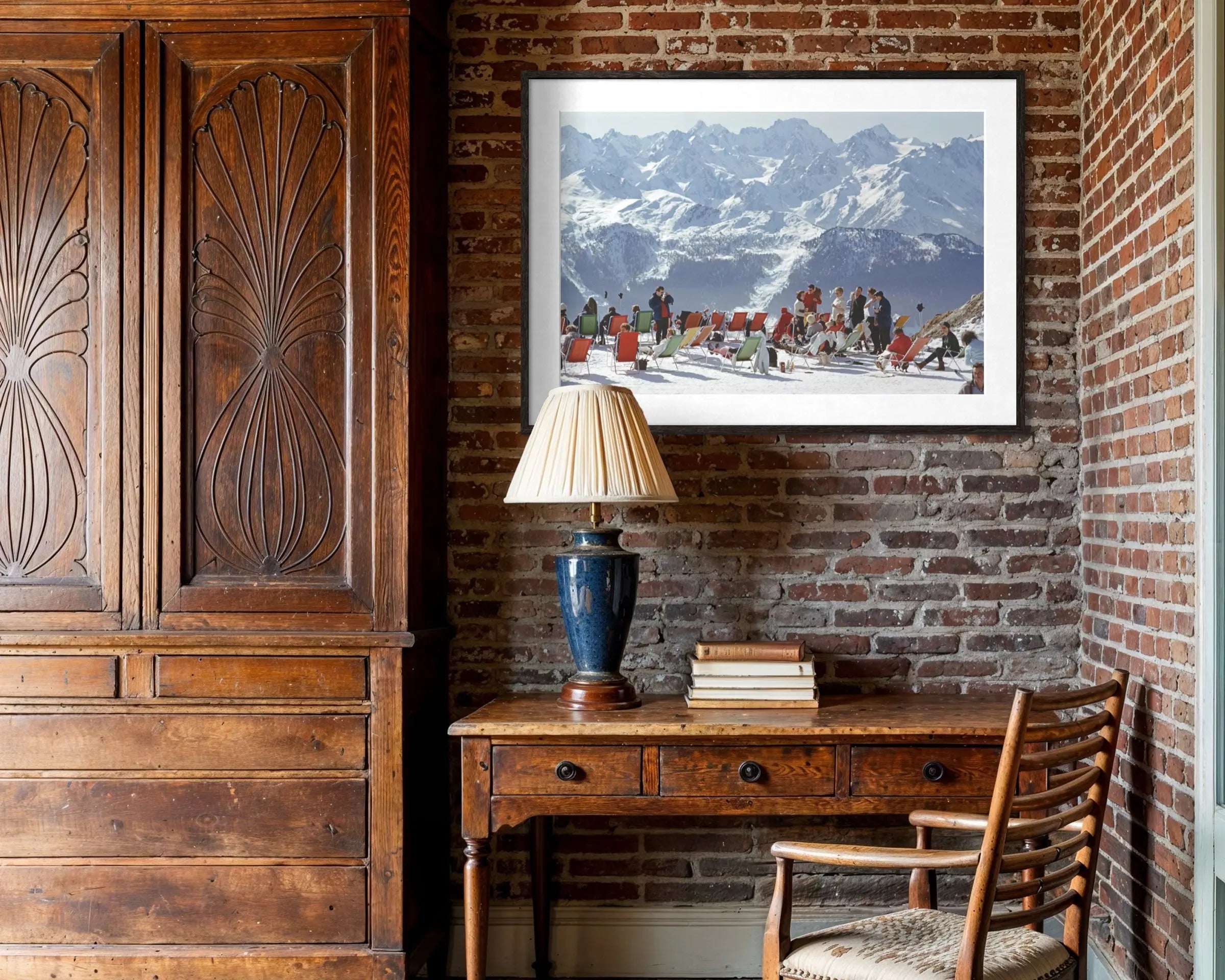 A framed art print depicting a snowy mountain landscape with figures in red clothing, displayed on a brick wall above a wooden desk in a rustic room setting.