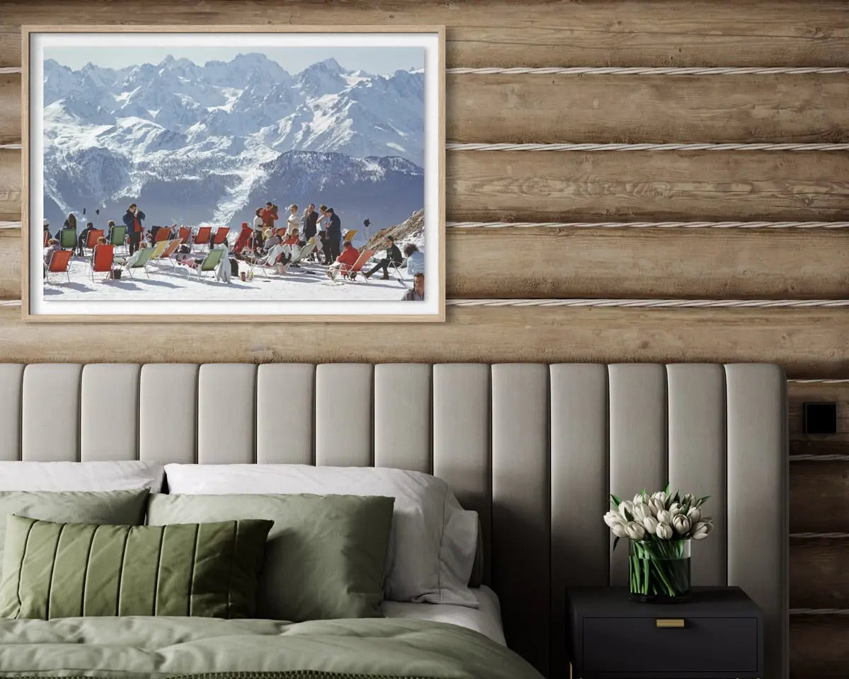 A framed art print depicting a snowy mountain ski resort with people, displayed on a rustic wood-paneled wall above a bed with green pillows and a bedside table.