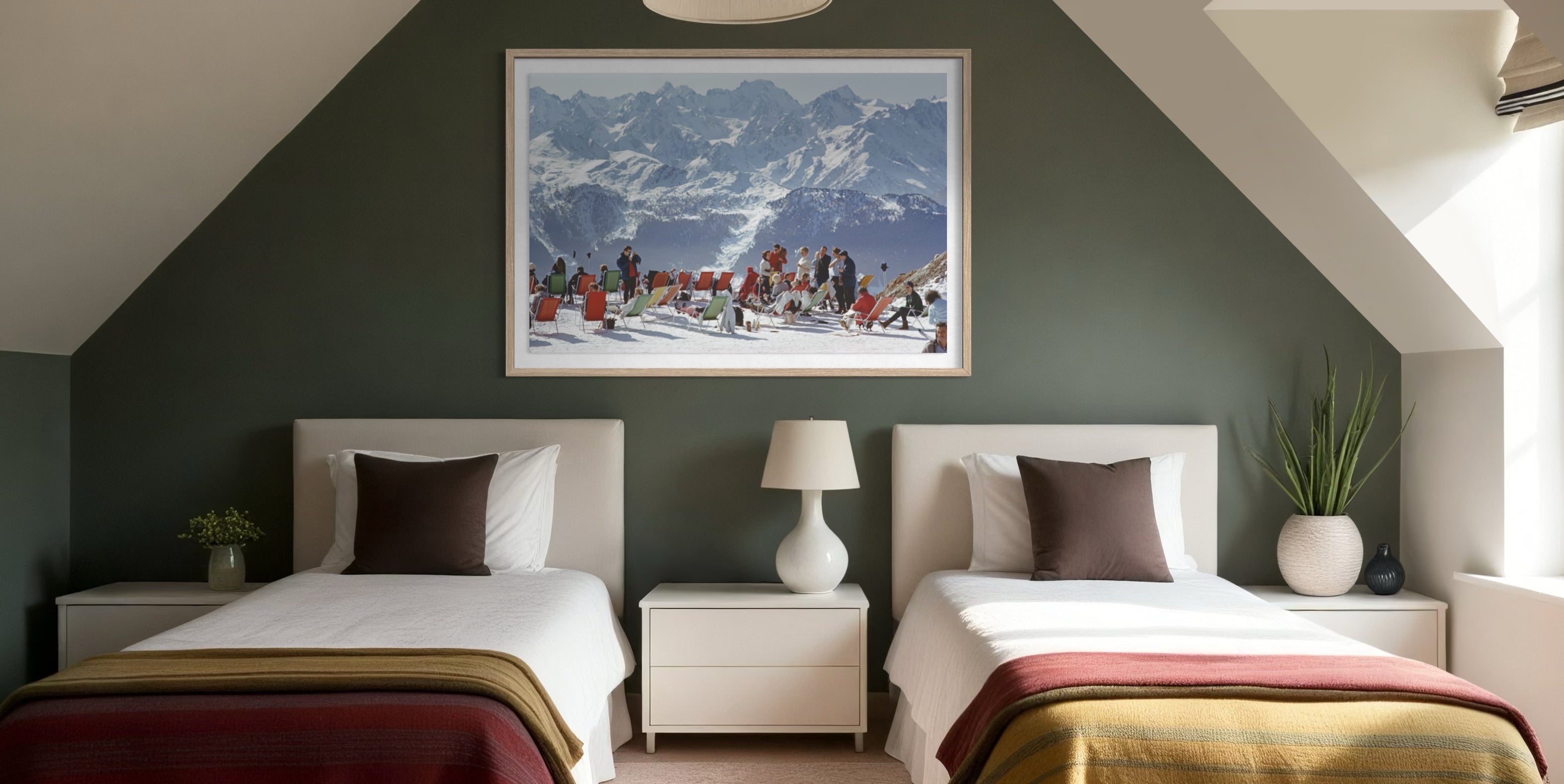 A framed art print depicting a snowy mountain ski resort with colorful skiers, displayed on a dark green wall above two single beds in a modern, symmetrically styled bedroom. The Picturalist Wall Art and Photography for Designed Interiors. Slim Aarons Collection, Skiing. Getty Images Gallery.