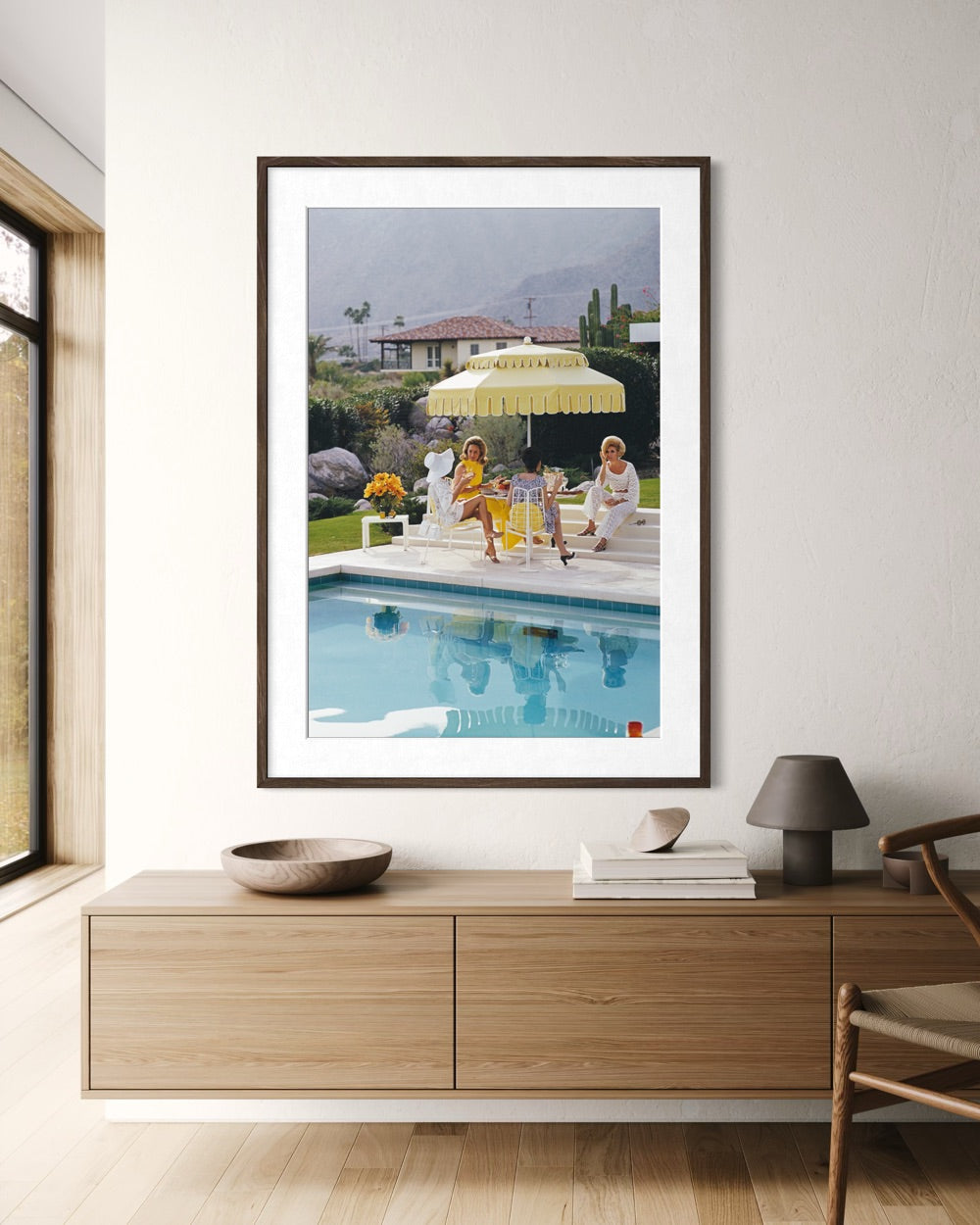 A framed art print depicting a sunny poolside scene with people under a yellow umbrella, displayed on a white wall above a modern wooden console table in a styled interior.