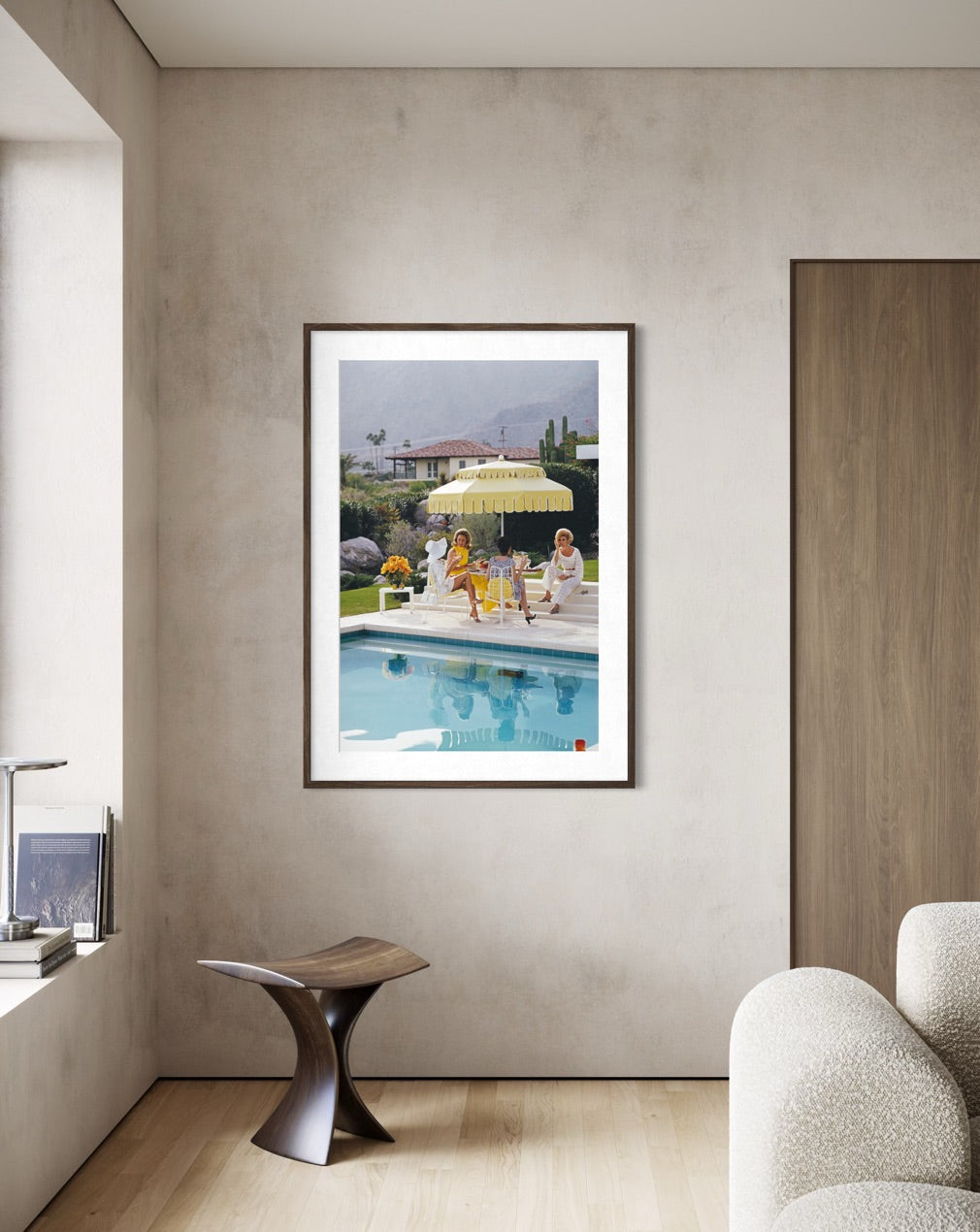 A framed art print depicting a sunny poolside scene with people under a yellow umbrella, displayed on a textured beige wall in a modern room setting.