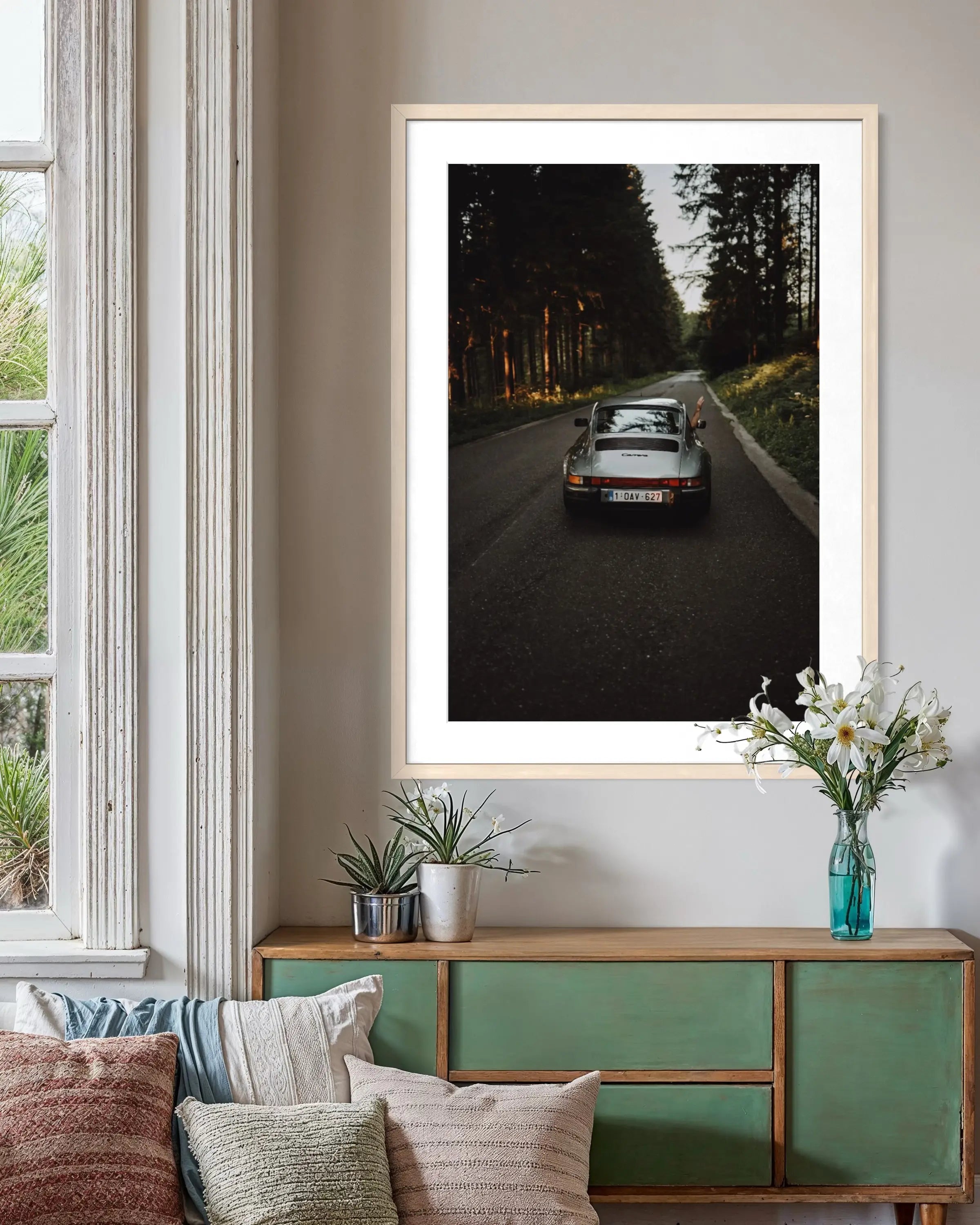 A framed vertical art print of a classic silver sports car driving down a dark forest road, displayed on a beige wall above a green wooden console table with decorative pillows and plants.