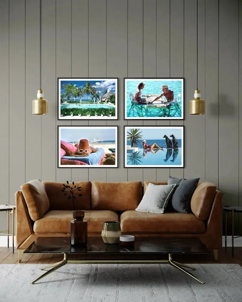 A set of four framed tropical beach and poolside art prints displayed on a gray wood-paneled wall above a brown leather sofa in a modern living room setting.