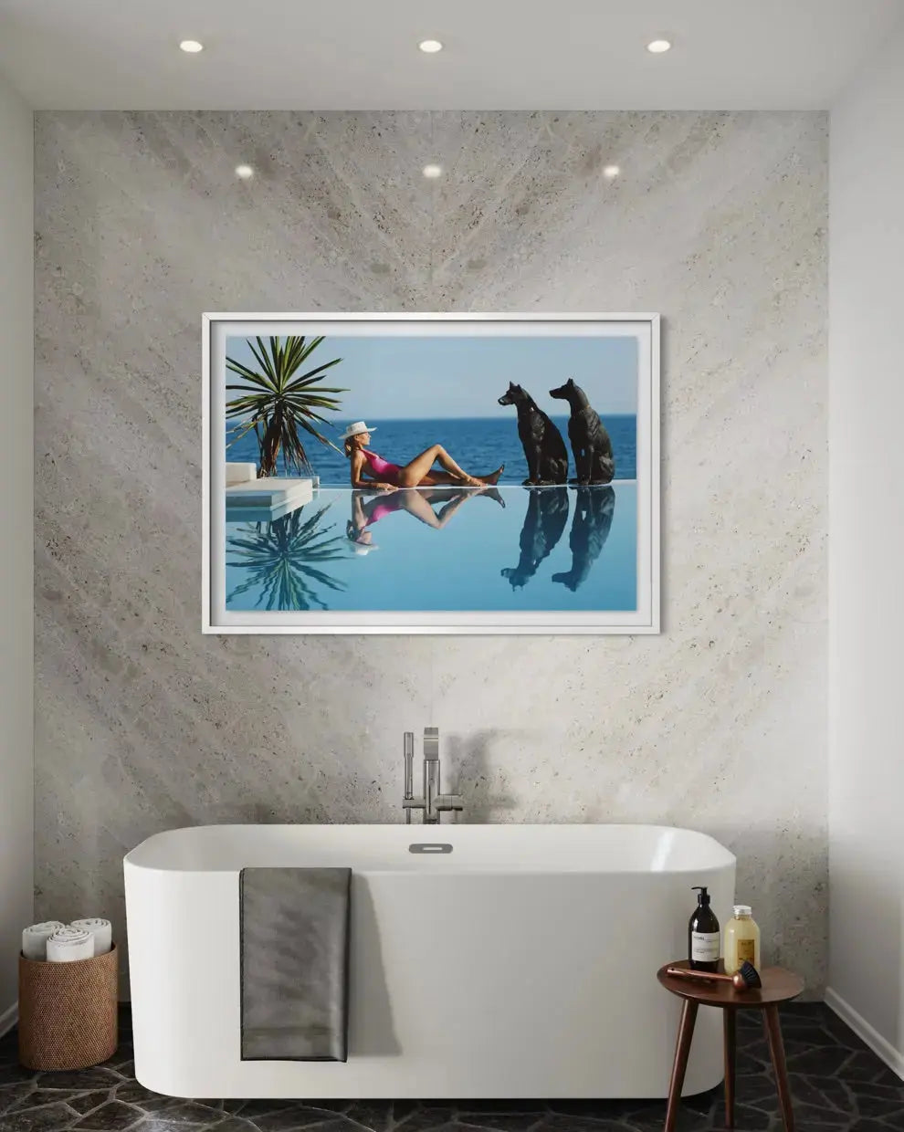 A framed art print depicting a woman lounging by an infinity pool with two dogs, displayed on a textured gray stone wall above a white freestanding bathtub in a modern bathroom setting.