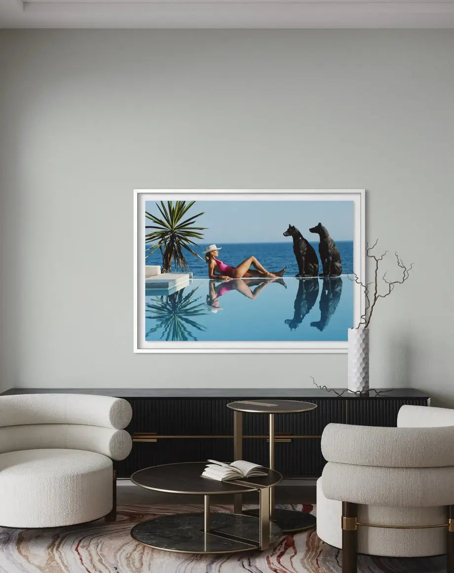 A framed art print depicting a woman relaxing by an infinity pool with two dogs, displayed on a light gray wall in a modern living room setting.