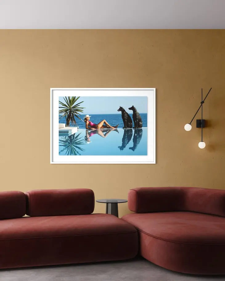 A framed art print depicting a woman relaxing by an infinity pool with two dogs, displayed on a warm beige wall above a modern red sectional sofa.