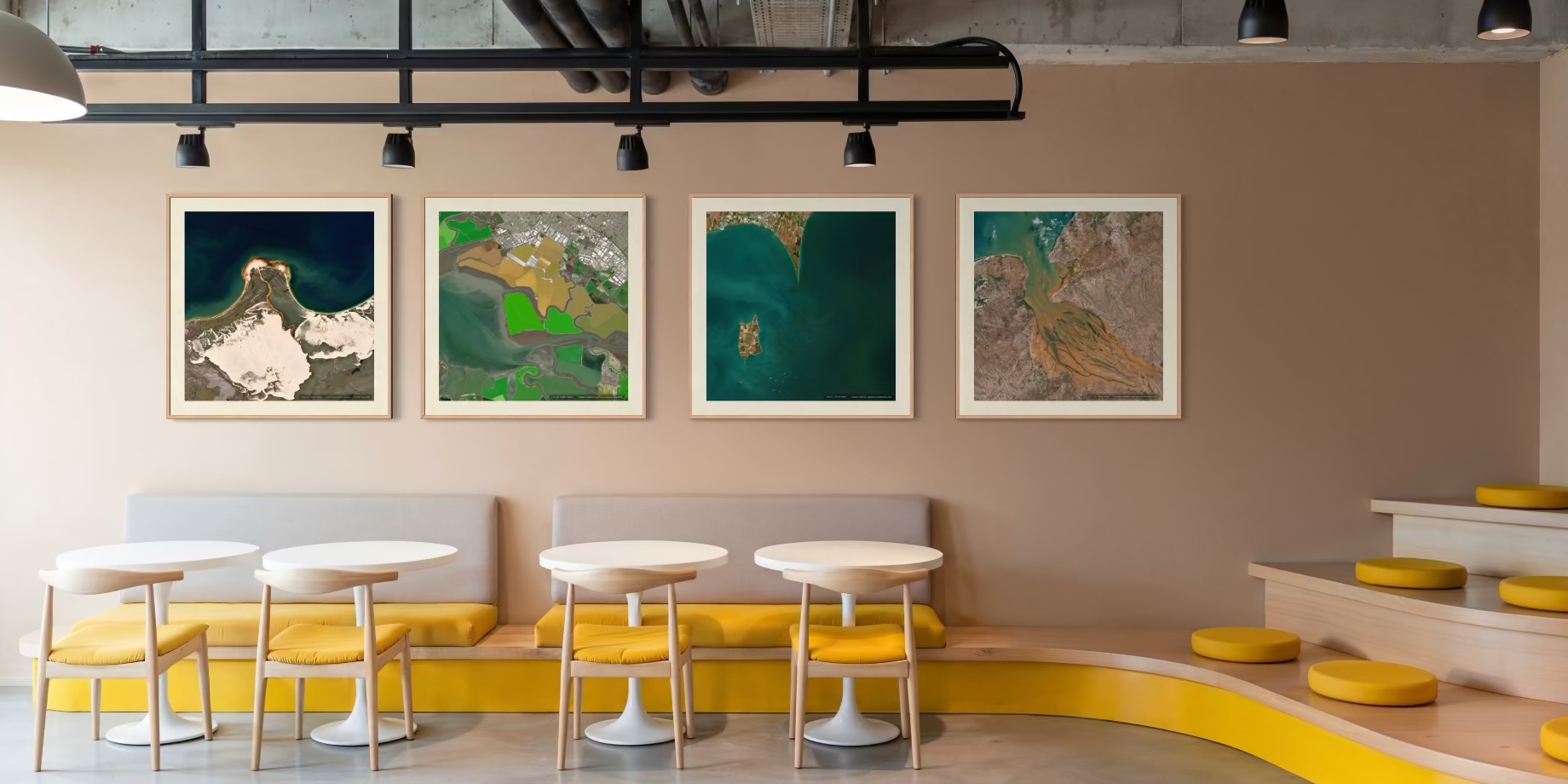 Four framed landscape art prints with white frames hang on a beige wall in a modern interior setting with curved yellow seating, white tables, and industrial ceiling details.