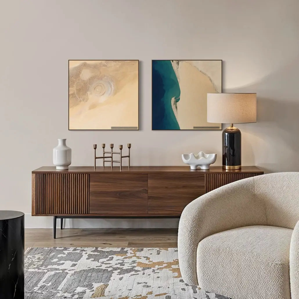 A styled interior scene showing two framed abstract wall art prints above a wooden sideboard, with a lamp, vase, candle holders, rug, and part of a cream upholstered chair in a living room setting.