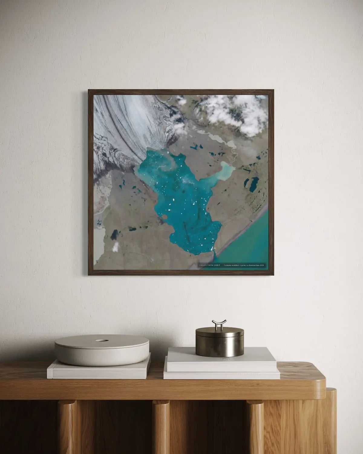 A framed art print featuring an aerial satellite view of a turquoise lake and surrounding mountains, displayed on a white wall above a wooden console table with books and decorative objects.