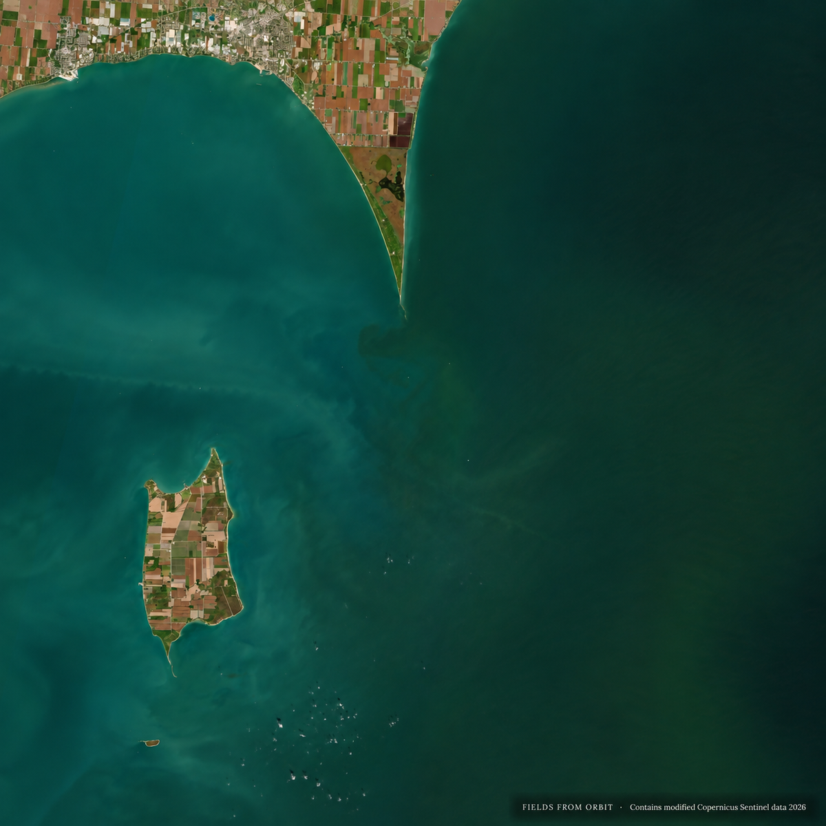 Satellite view of a coastal landscape with a small peninsula on the left, a large body of deep blue-green water on the right, and a narrow strip of shoreline at the top edge.