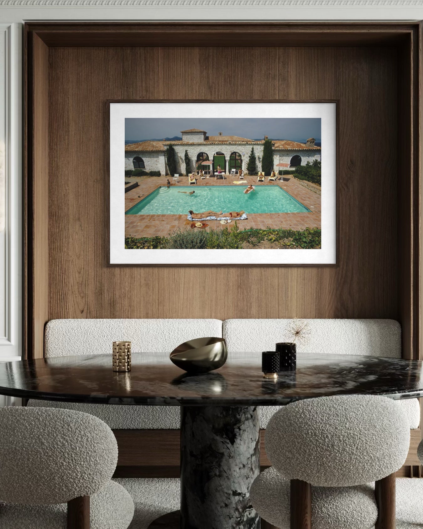 A framed art print depicting a luxurious outdoor swimming pool and villa courtyard, displayed on a dark wood wall above a dining table with textured chairs.