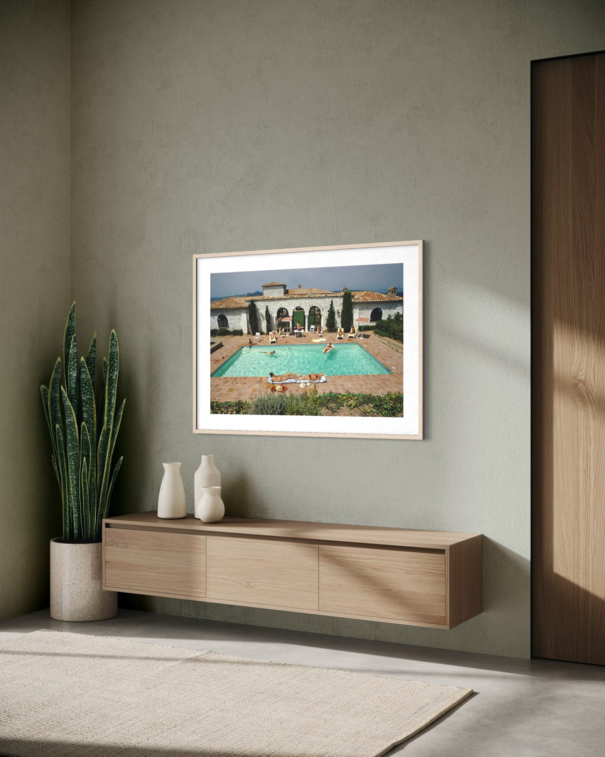 A framed art print depicting a Mediterranean-style swimming pool courtyard with a villa, displayed on a textured sage green wall above a floating wooden console table in a modern living room setting.