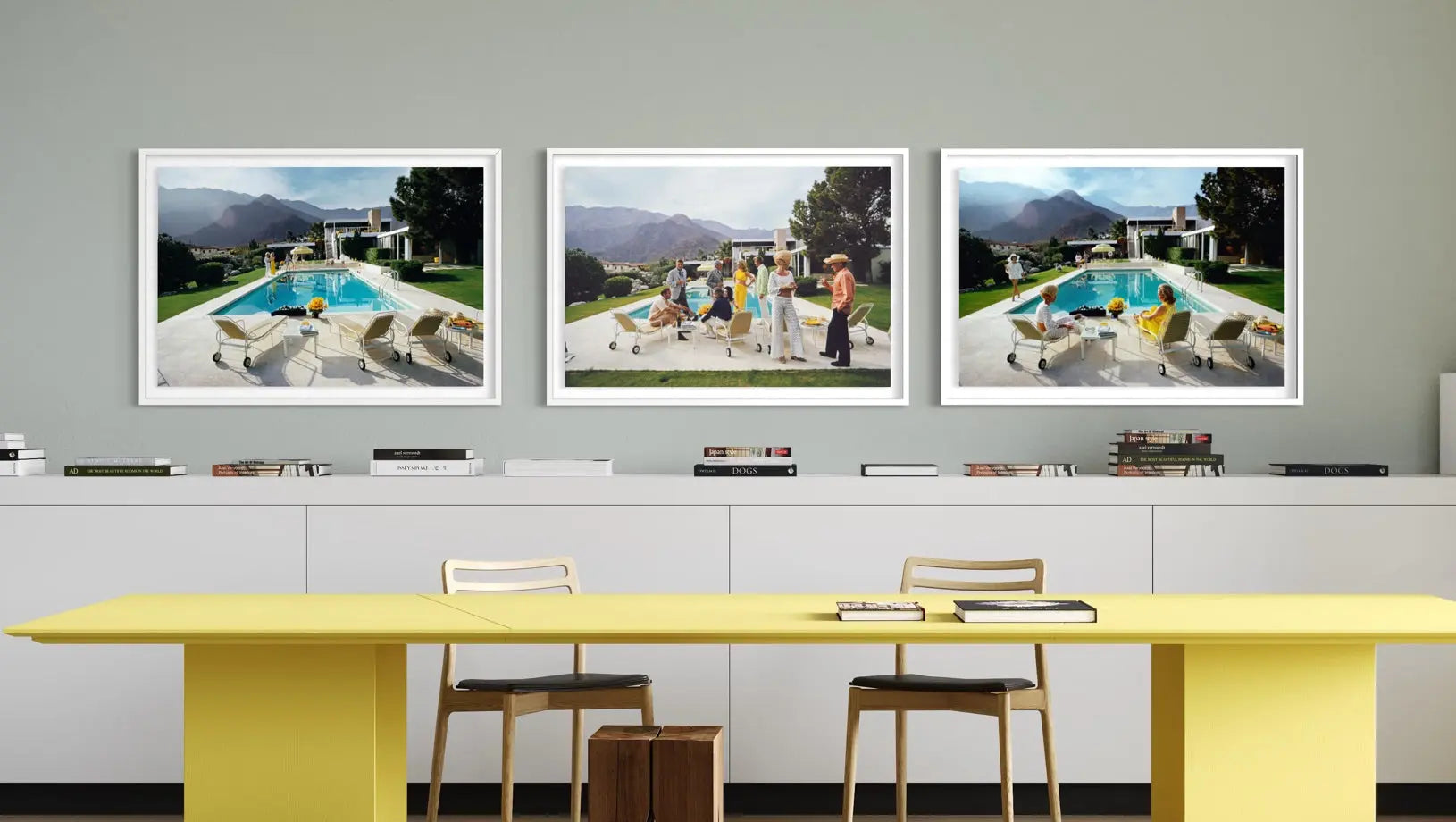 A set of three framed art prints depicting outdoor poolside scenes, displayed on a gray wall above a modern yellow table and white chairs. The Picturalist Wall Art and Photography for Designed Interiors. Slim Aarons Collection, Poolside. Getty Images Gallery.