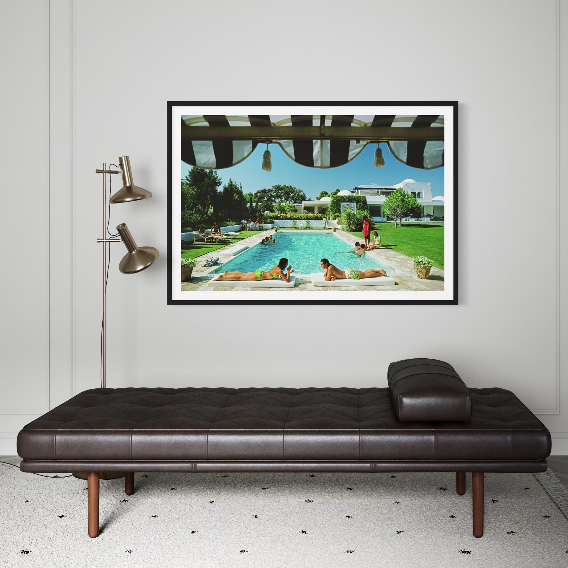 Framed artwork of people by a pool on a wall above a brown leather daybed.
