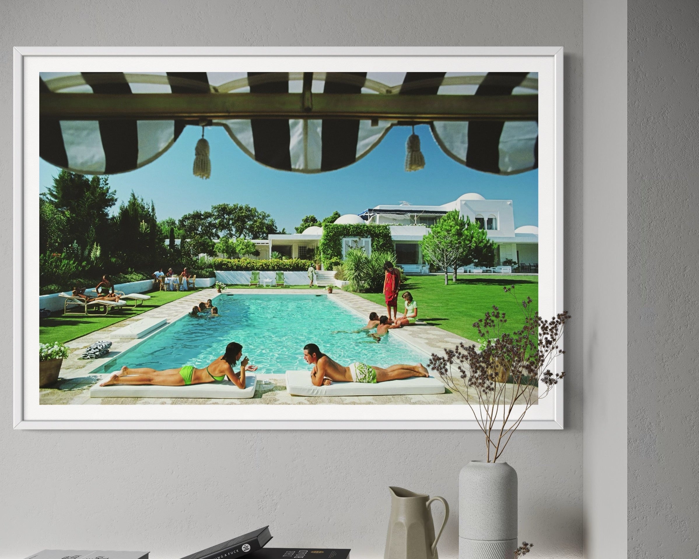 Framed photograph of a pool scene with people lounging, displayed on a wall above a shelf.