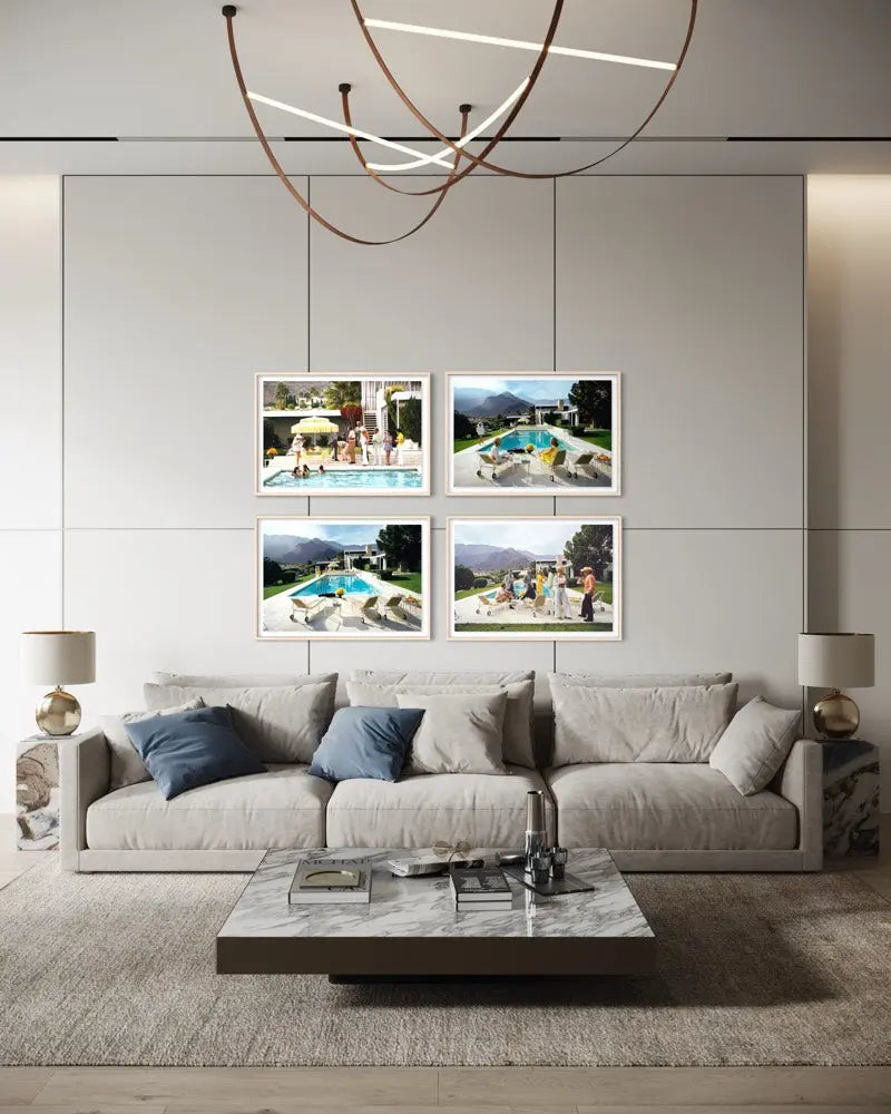A modern living room with a light beige sofa, marble coffee table, and a wall-mounted set of four framed photographs above the couch. The photos depict outdoor poolside and mountain scenes. The Picturalist Wall Art and Photography for Designed Interiors. Slim Aarons Collection, Poolside. Getty Images Gallery.