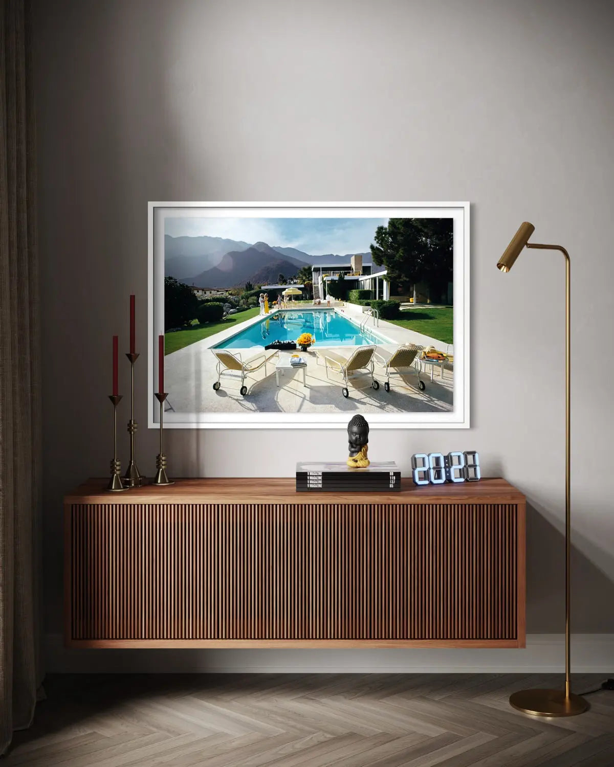 A framed art print depicting a luxurious outdoor poolside scene with mountains in the background, displayed on a gray wall above a wooden console table in a modern living room setting. The Picturalist Wall Art and Photography for Designed Interiors. Slim Aarons Collection, Poolside. Getty Images Gallery.