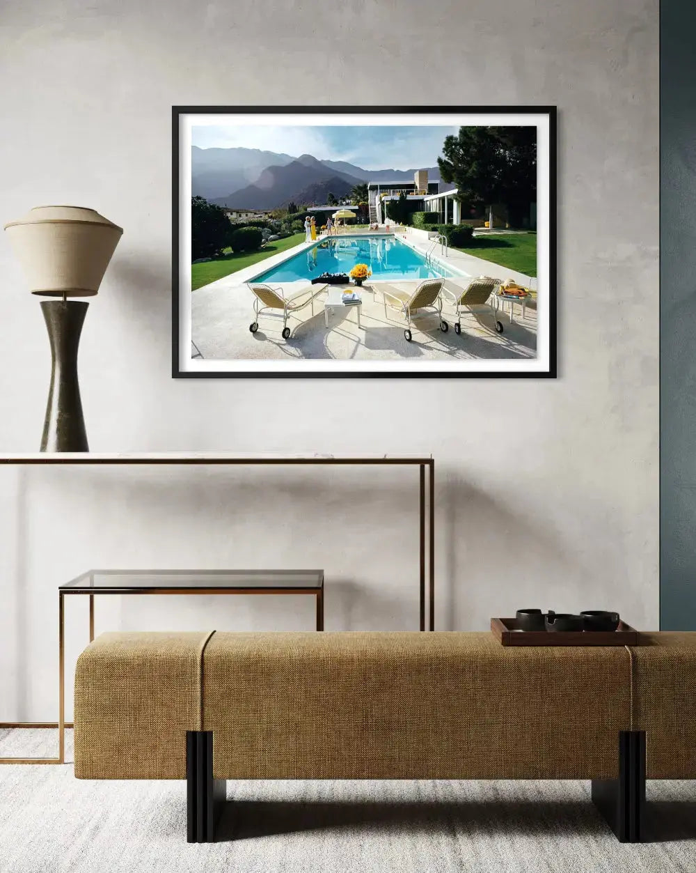 A framed art print depicting a luxurious outdoor poolside scene with mountains in the background, displayed on a textured light gray wall above a modern console table and bench. The Picturalist Wall Art and Photography for Designed Interiors. Slim Aarons Collection, Poolside. Getty Images Gallery.