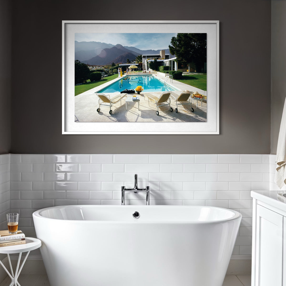 A framed art print depicting a luxurious outdoor poolside scene with lounge chairs, mountains in the background, and a modern house, displayed on a dark gray wall above a white tiled bathroom wall and freestanding bathtub.