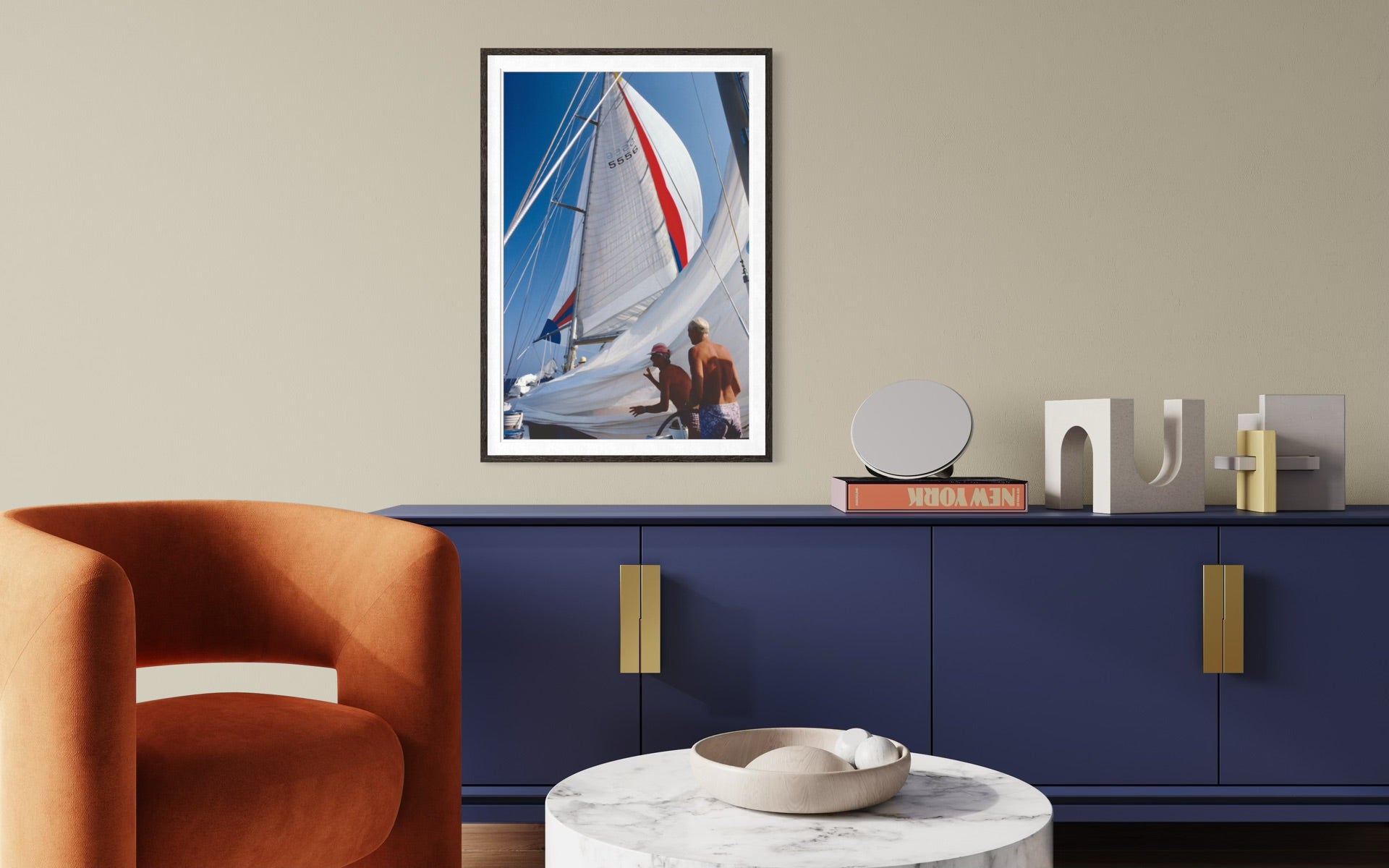 A framed vertical art print of a sailboat on blue water, displayed on a beige wall above a navy blue cabinet in a modern living room setting.