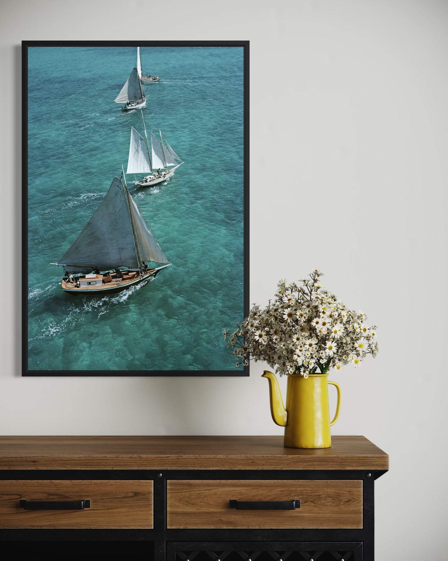 Framed artwork of sailboats on a wall above a wooden console table with a yellow pitcher and flowers.