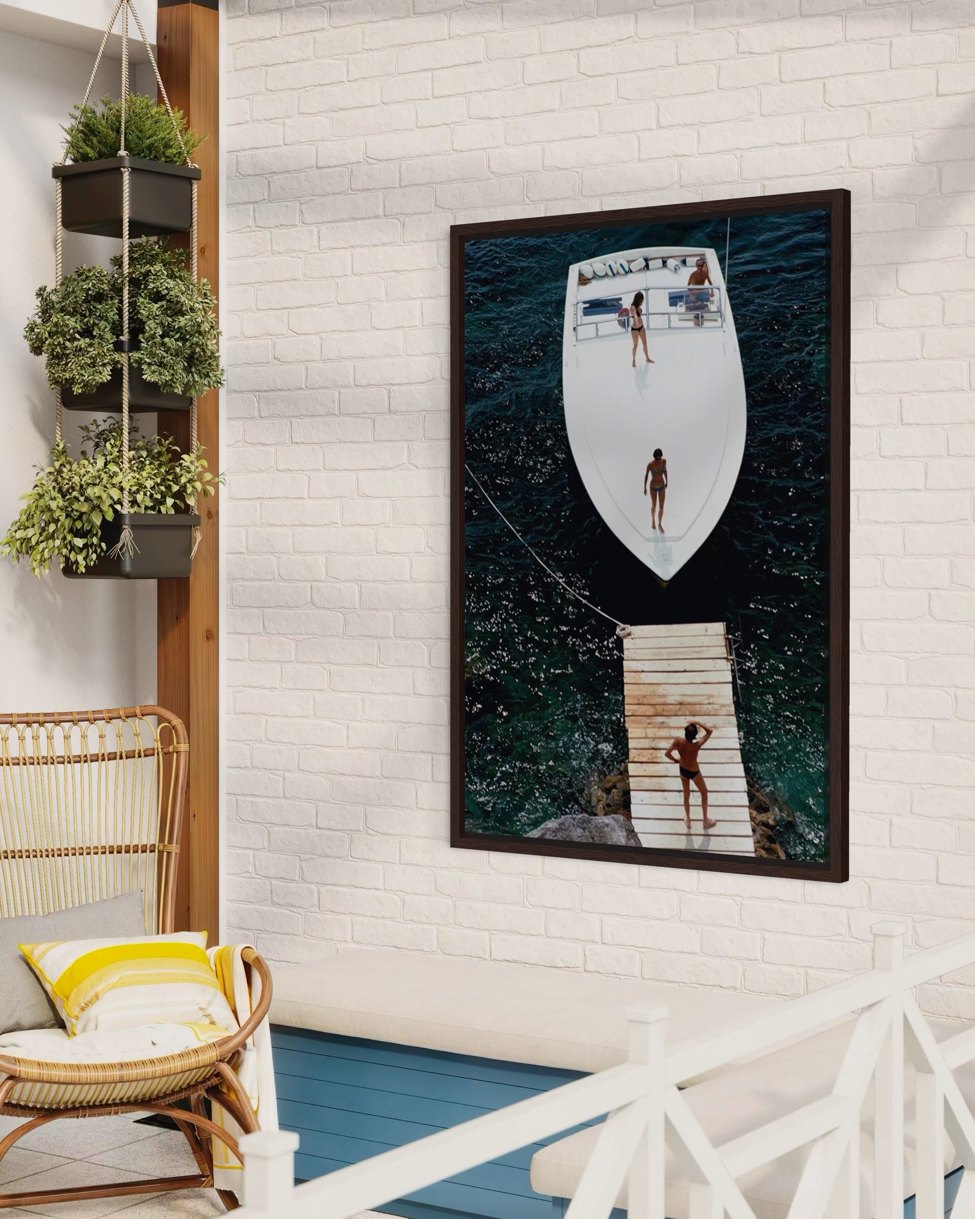 Framed artwork of a boat on a white brick wall with a wicker chair and plants below.