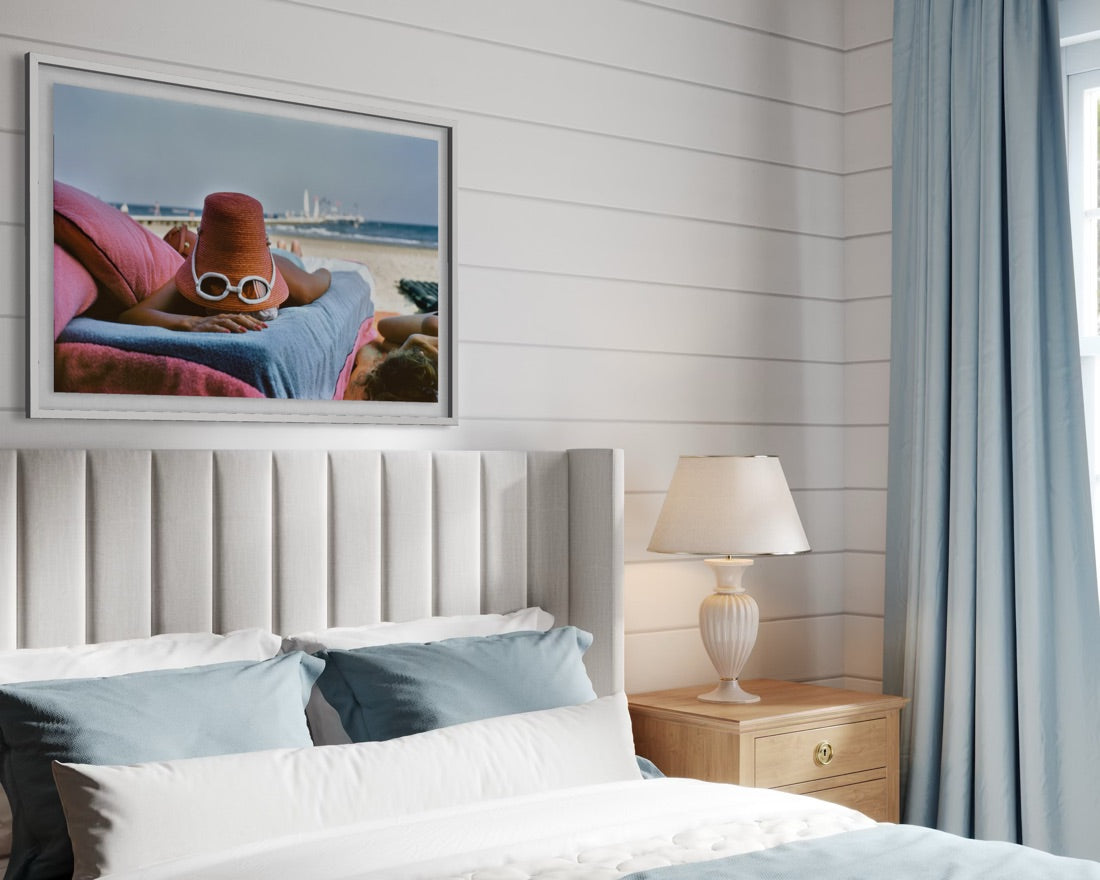 A framed art print depicting a person lounging on a pink beach chair by the ocean, styled in a bedroom above a bed with white and blue bedding, a wooden nightstand with a lamp, and light blue curtains.