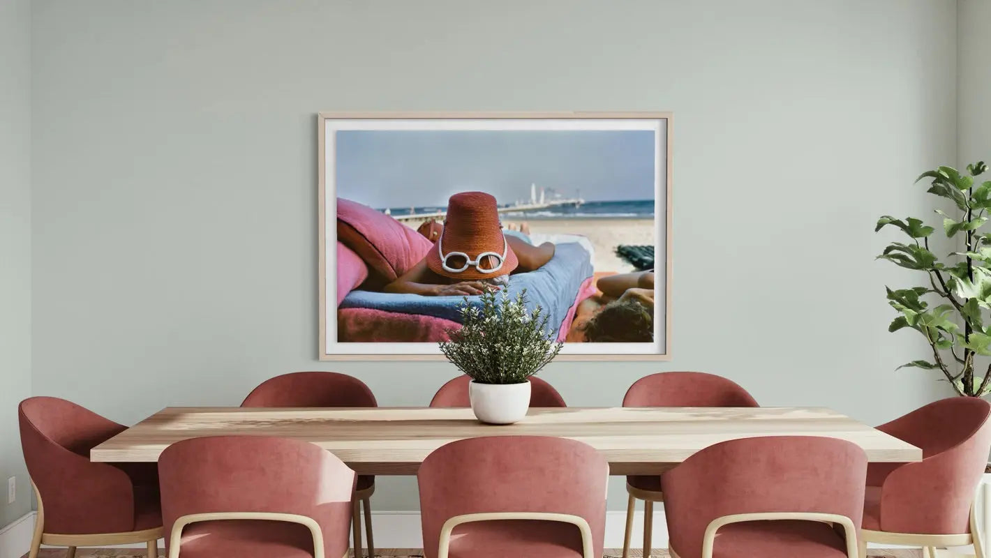 A framed art print depicting a woman lounging on a beach lounge chair by the ocean, hung on a light gray wall above a wooden dining table with pink chairs.