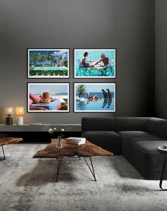 A modern living room setting featuring a four-panel framed wall art set depicting tropical beach and poolside scenes, mounted on a dark gray wall above a contemporary dark gray sectional sofa and a rustic wood coffee table.