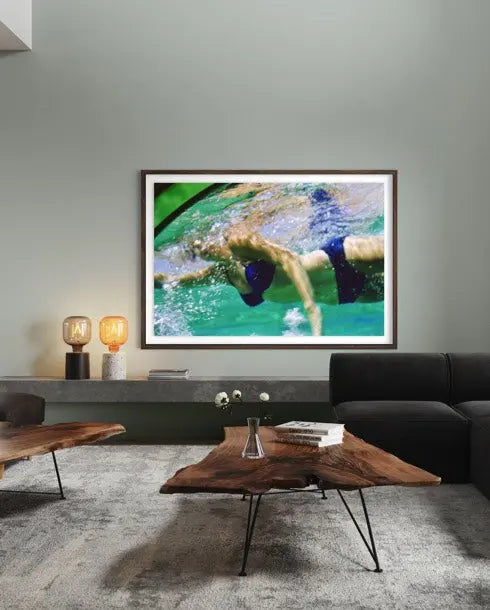 A framed art print depicting a swimmer in turquoise water, displayed on a gray wall in a modern living room setting above a wooden coffee table and dark sofa.