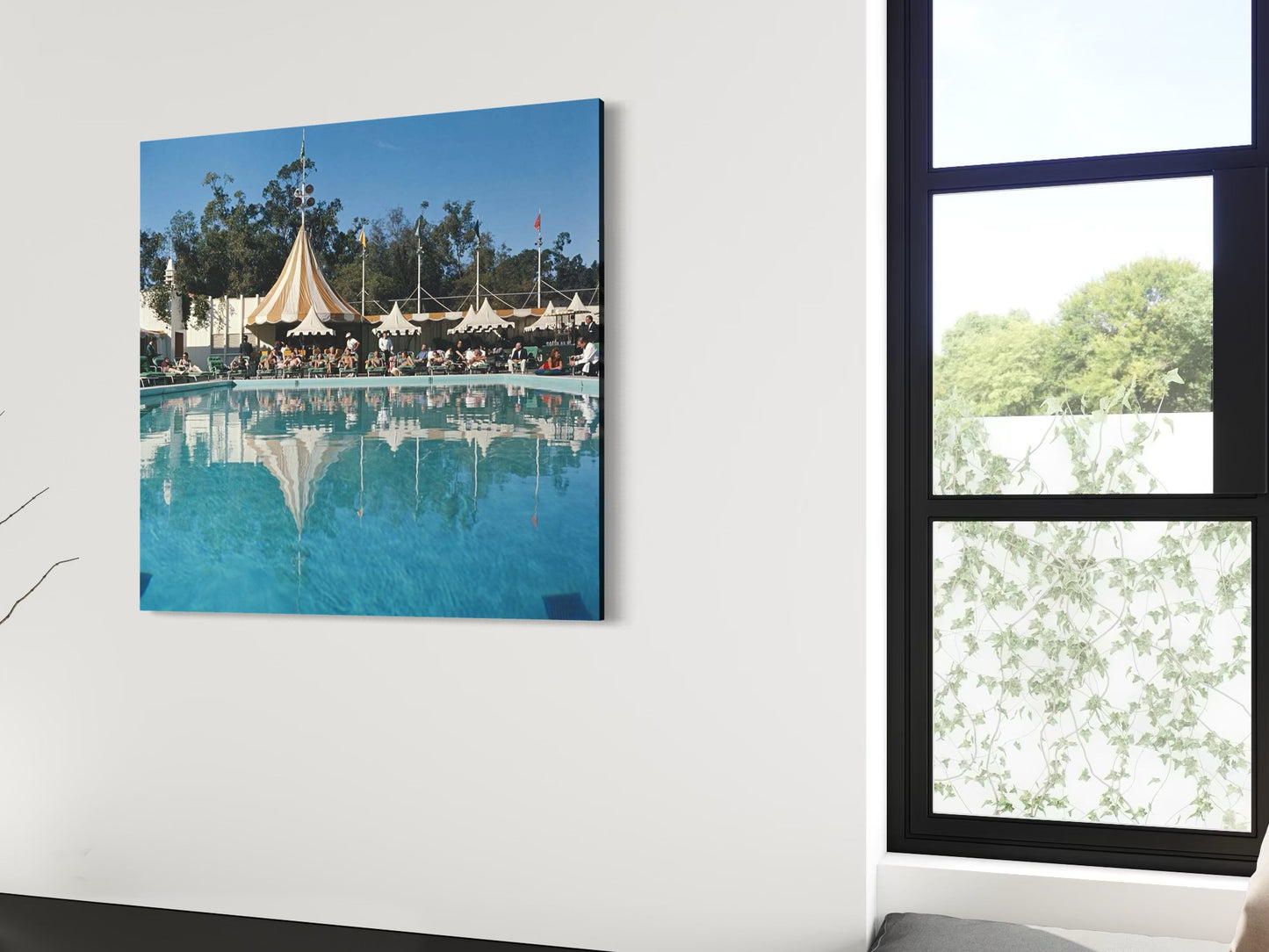 Unframed metal print of a pool scene with tents on a white wall next to a window.