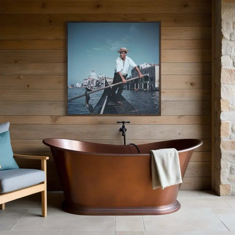 A framed art print depicting a gondolier in a gondola in Venice, hung on a wooden plank wall above a freestanding copper bathtub. The Picturalist Wall Art and Photography for Designed Interiors. Slim Aarons Collection, Venice. Getty Images Gallery.