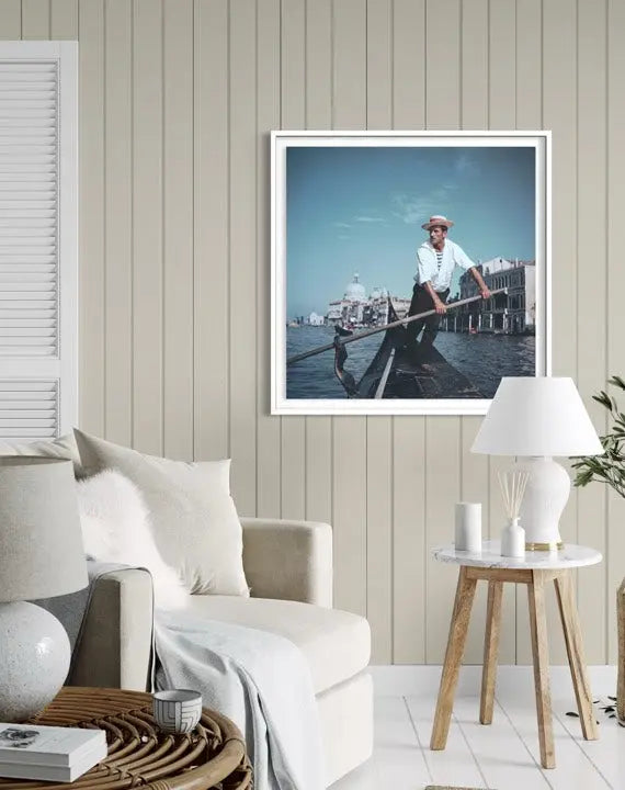 A framed art print depicting a man rowing a gondola in Venice, displayed on a beige wood-paneled wall in a modern, neutral-toned living room setting. The Picturalist Wall Art and Photography for Designed Interiors. Slim Aarons Collection,Venice. Travel. Getty Images Gallery.