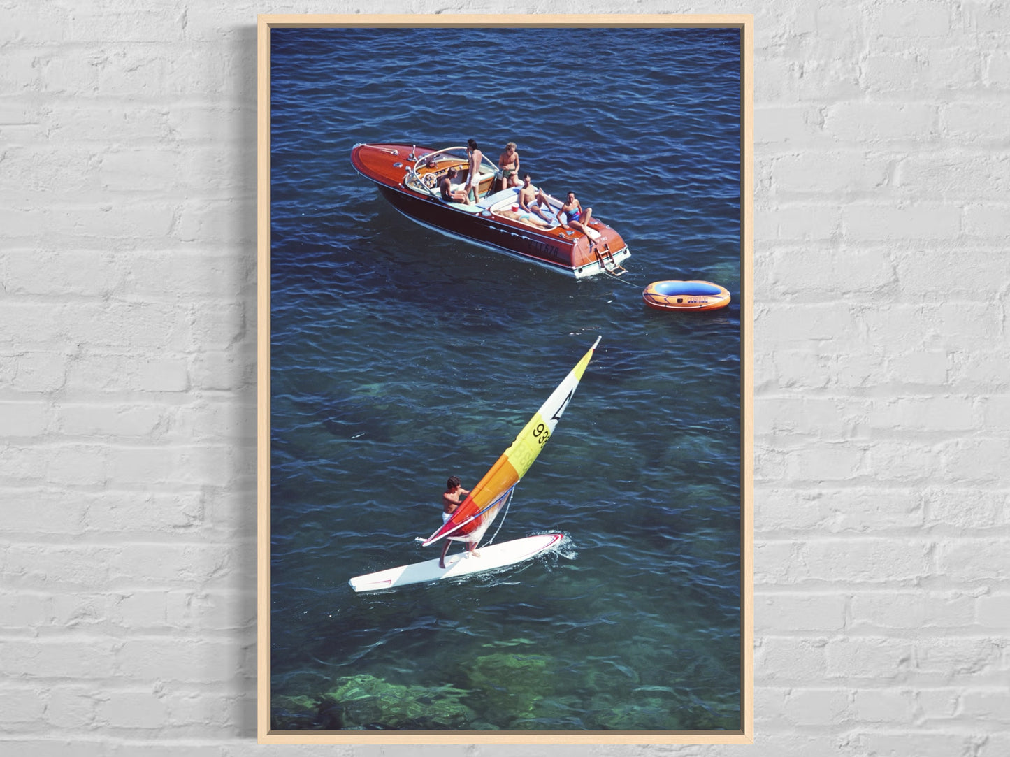 Framed artwork of a boat and sailboard on water against a brick wall.