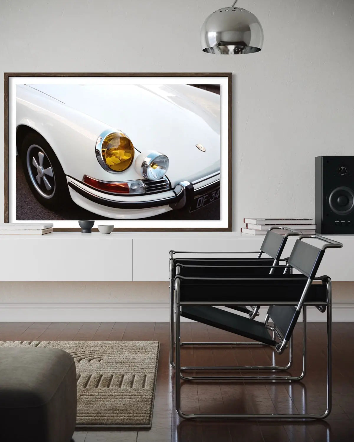 A framed wall art print showing a close-up photograph of a white classic car, displayed in a modern living room setting above a white console table with two black chairs and a speaker.