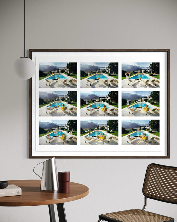 A framed wall art print featuring a 3x3 grid of nine resort pool scenes with mountains in the background, displayed on a light gray wall above a wooden table with a lamp and chair. The Picturalist Wall Art and Photography for Designed Interiors. Slim Aarons Collection, Poolside. Getty Images Gallery.